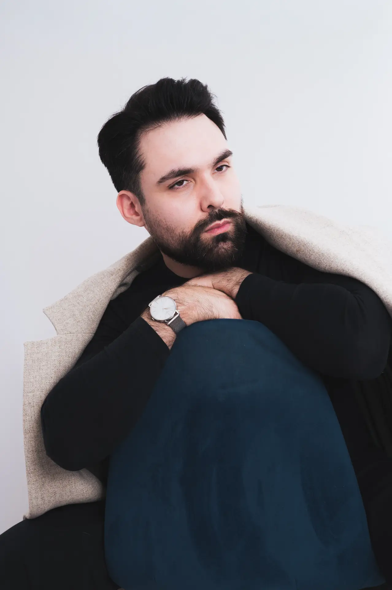 Homme en pull noir, mains sous le menton, dans un studio photo fond blanc sur une chaise bleu